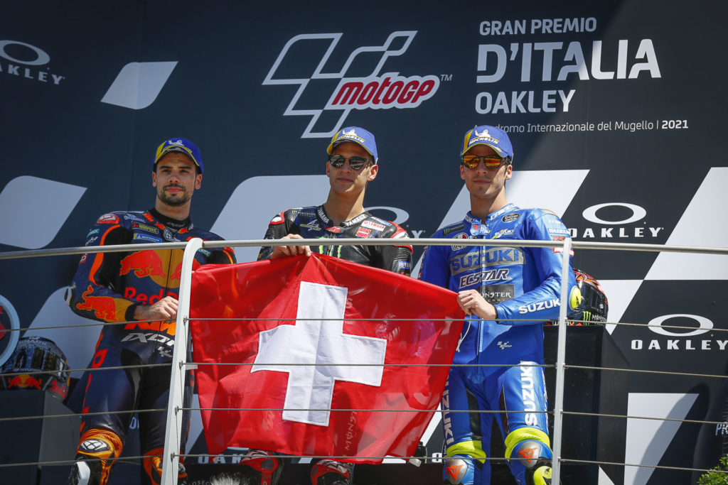 MotoGP race winner Fabio Quartararo (center), runner-up Miguel Oliveira (left), and third-place finisher Joan Mir (right) on the podium holding a Swiss flag in honor of fallen Moto3 racer Jason Dupasquier. Photo courtesy Dorna.