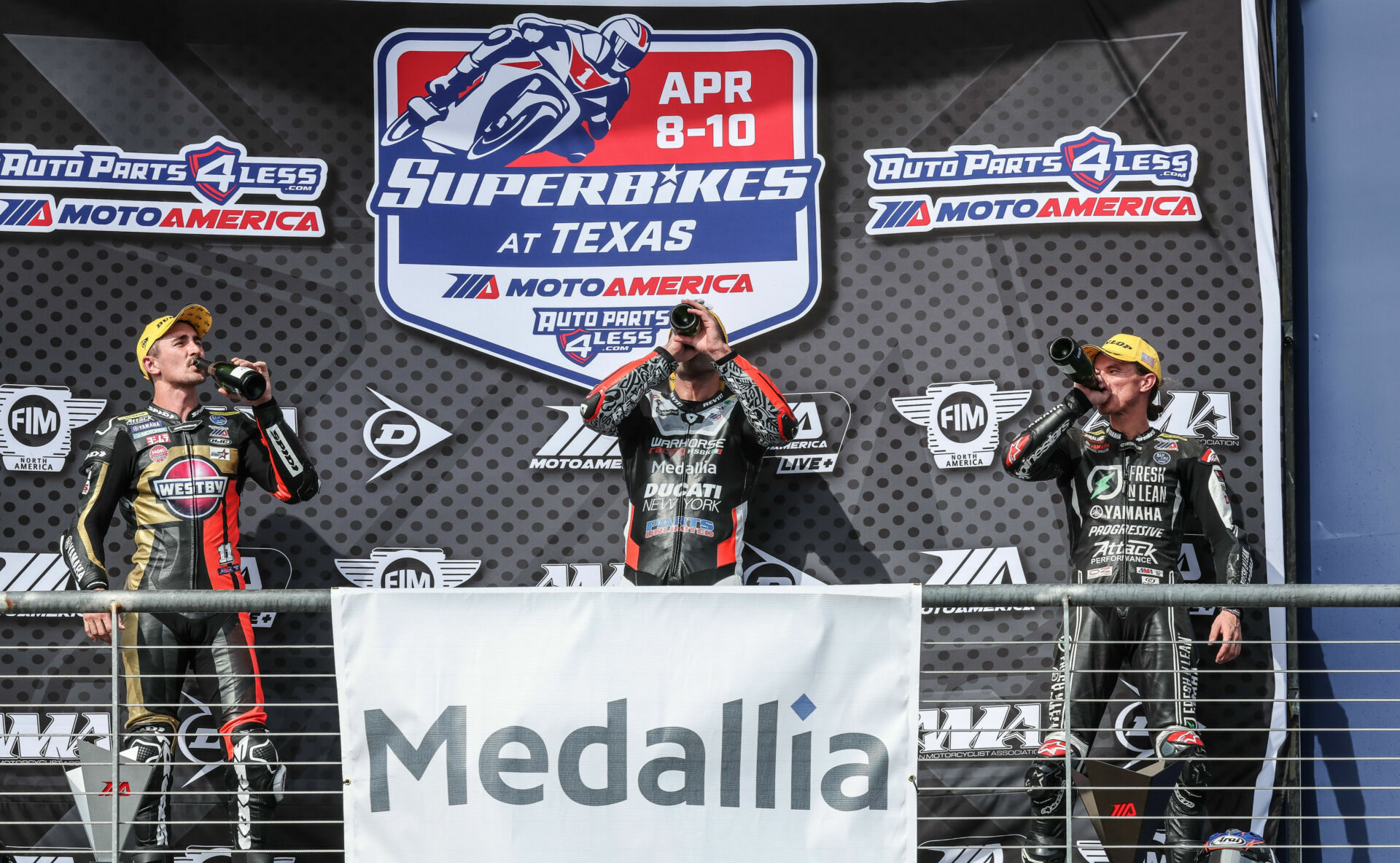 MotoAmerica Medallia Superbike Race Two winner Danilo Petrucci (center), runner-up Mathew Scholtz (left), and third-place finisher Jake Gagne (right) on the podium at COTA with Auto Parts 4 Less logos displayed prominently on the podium backdrop. Photo by Brian J. Nelson.
