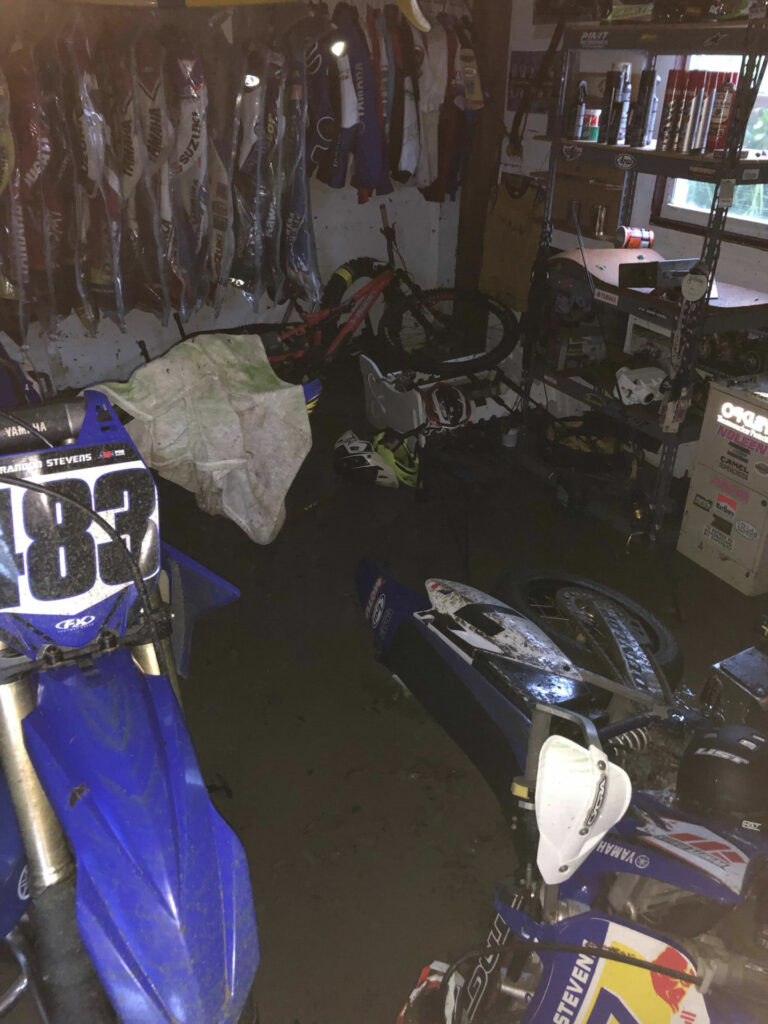 Thomas Stevens' garage/basement after Hurricane Ian with his supermotard lying on its side in sea water, his Yamaha YZ250 motocrosser standing upright next to it, and his old race leathers hanging in the background. Photo courtesy Thomas Stevens.