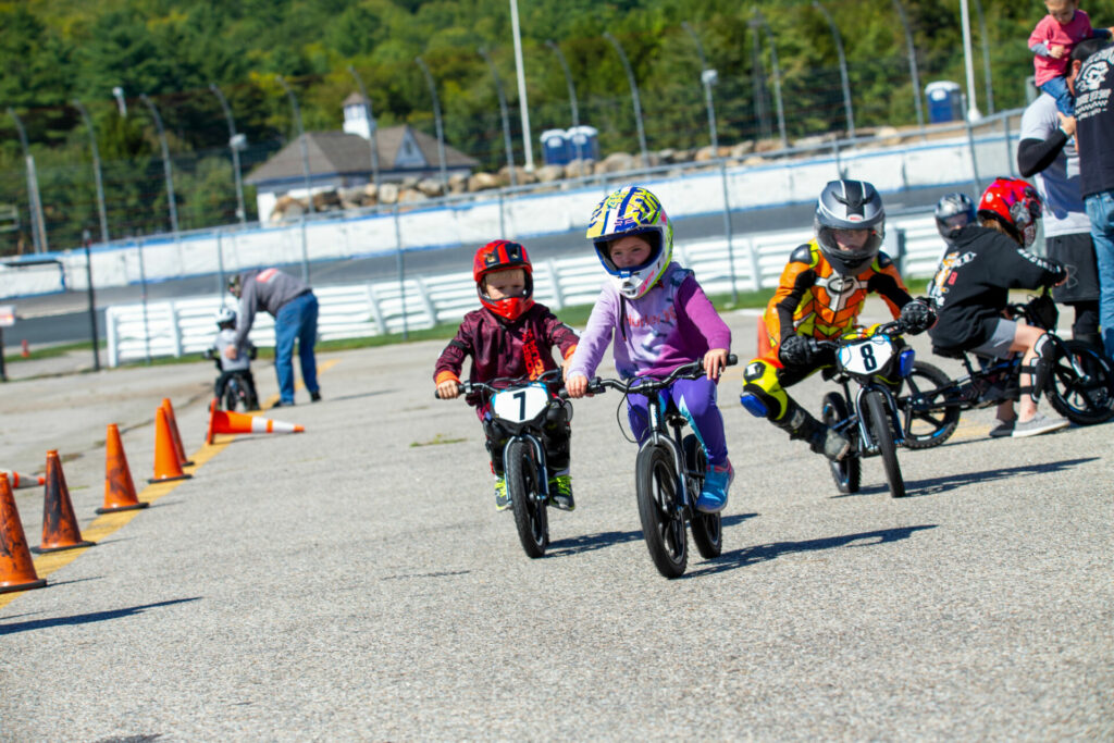 Kids of varying skill levels riding StaCyc electric balance bikes while a volunteer works with another rider in the background. Photo by Sam Draiss, courtesy NEMRR.