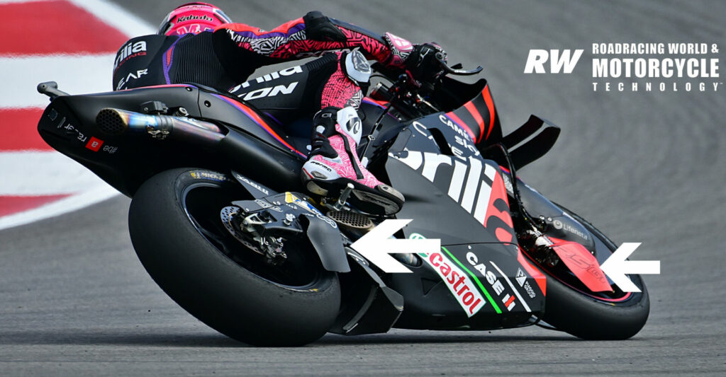Fairing And Swingarm: Aprilia continues to use the “fat” fairing lower to create downforce and clean and direct airflow under the machine when it is on its side in a corner. The manufacturer has gone a step further with wings bolted directly to the swingarm and front wheel fender (white arrows) that apply downforce directly to the tire, instead of transmitting it through the suspension as fairing-mounted aerodynamic devices do. Photo by Michael Gougis.