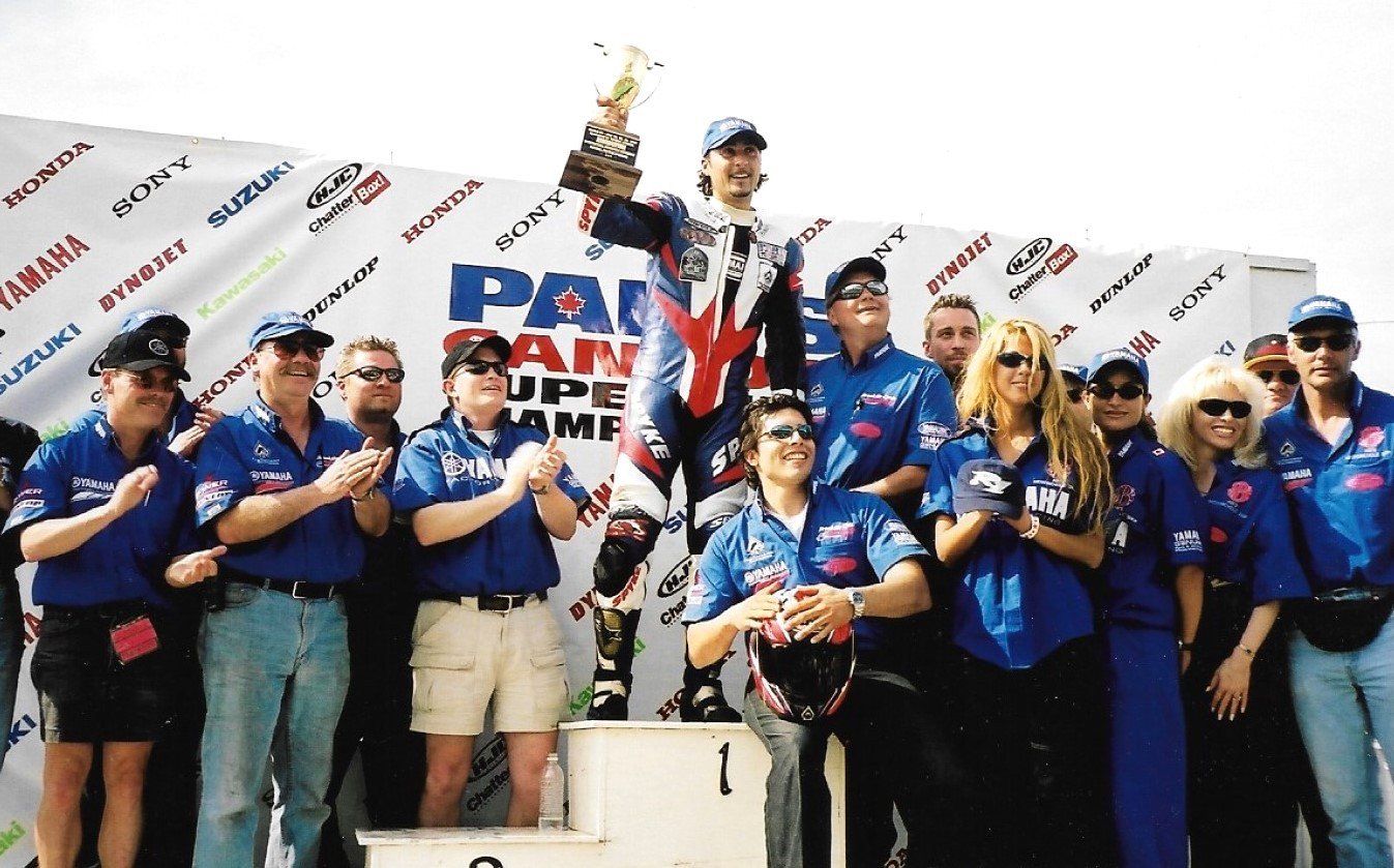 2002 Parts Canada Superbike Pro Open Sport Bike NationalChampionship podium at Shannonville Motorsport Park (class of 2025) features
new number one Frank Trombino (centre) as well as bike builder Rob Egan of
Brooklin Cycle Racing (Class of 2025, glasses and hat, on the right of the
"war lord of Shannonville") and some of the staff of Ab's Cycle, another
2025 Canadian Motorcycle Hall of Fame inductee. Photo by Colin Fraser.